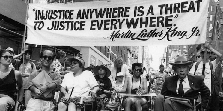 A black and white photo of a protest with a banner above a group of disabled activists