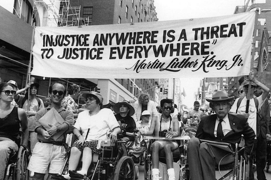 A black and white photo of a protest with a banner above a group of disabled activists