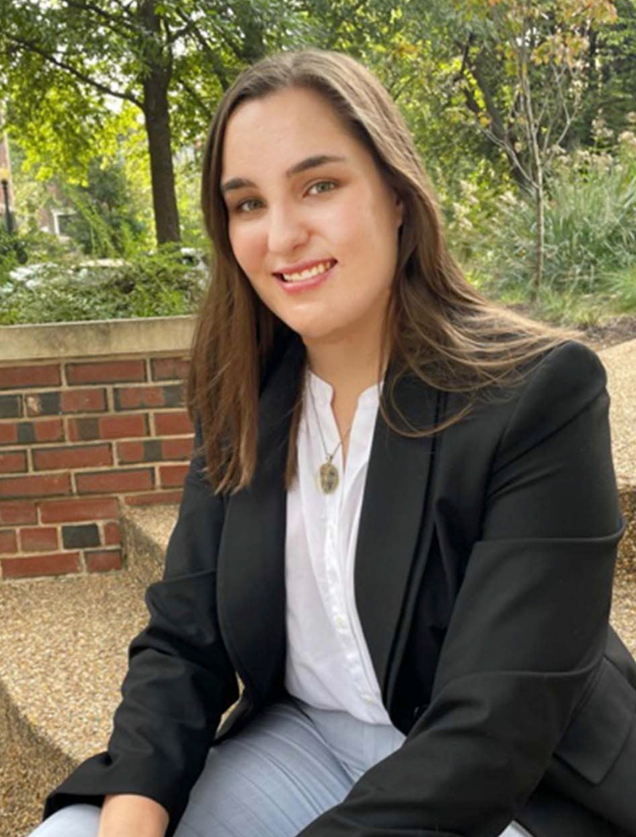 A photo of Rebecca sitting on the stairs in front of a brick wall with trees. She is a white woman with long brown hair. She is wearing a black blazer with a white top and blue pants