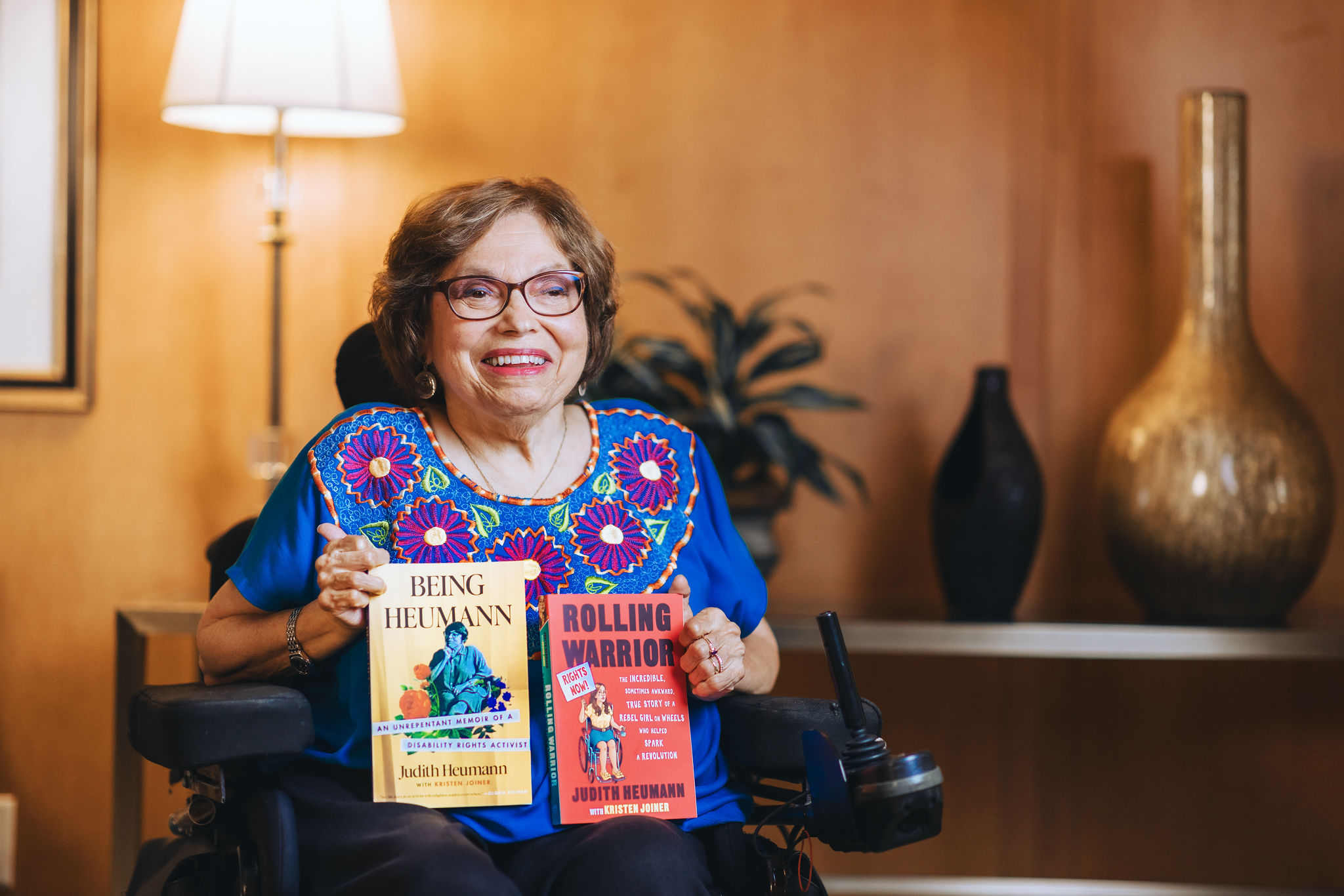 Photograph of Judith Heumann posing in her wheelchair holding her two books, Being Heumann and Rolling Warrior