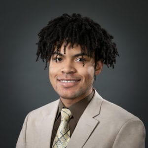 A Black man with short dreadlocks smiling at the camera and wearing a tan colored suit with a matching tie