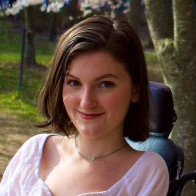 A white woman with chin length straight black hair. She uses a motorized wheelchair and is wearing a pink top. She is in front of some greenery.