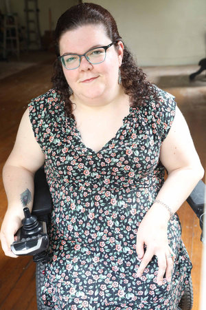 Emily Ladau is a white woman with long curly brown hair. She uses a motorized wheelchair and is wearing a floral dress.