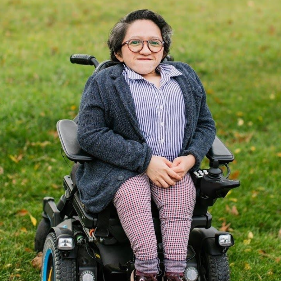 A photo of Sandy Ho. Sandy is an Asian American woman with short black hair. Sandy uses a wheelchair and is in a patch of grass. Sandy is wearing a jacket overtop of a collared shirt with khaki pants and black sneakers