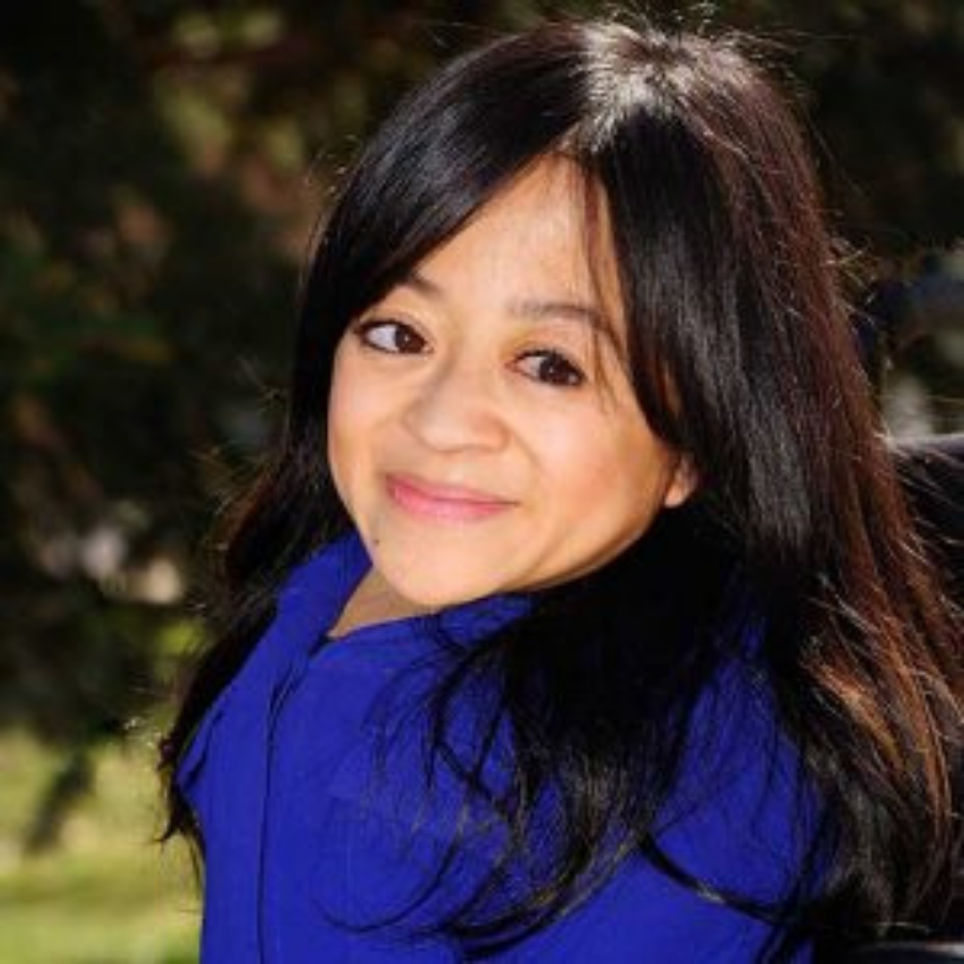 A headshot of Teresa Nyugen. Teresa is an Asian American woman who uses a wheelchair. She has long dark brown wavy hair. She is wearing a royal blue top and is smiling