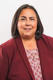 A headshot of Kathy Martinez, who is Latina and has shoulder length brown hair. She is wearing a red blazer with a pink patterned short underneath.