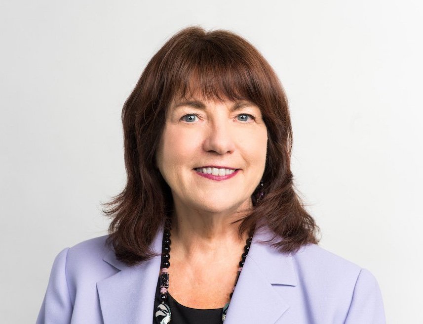 A headshot of Susan Reinhard who is a white woman with reddish-brown shoulder length hair. She is smiling and wearing a lilac blazer and black patterned top. There is a white background behind her.