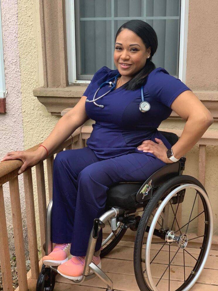 Andrea is a black woman who uses a manual wheelchair. She is pictured outdoors in front of a window. She is wearing navy blue nursing scrubs and pink shoes. Her black hair is in a ponytail and she has a stethoscope hanging around her neck.