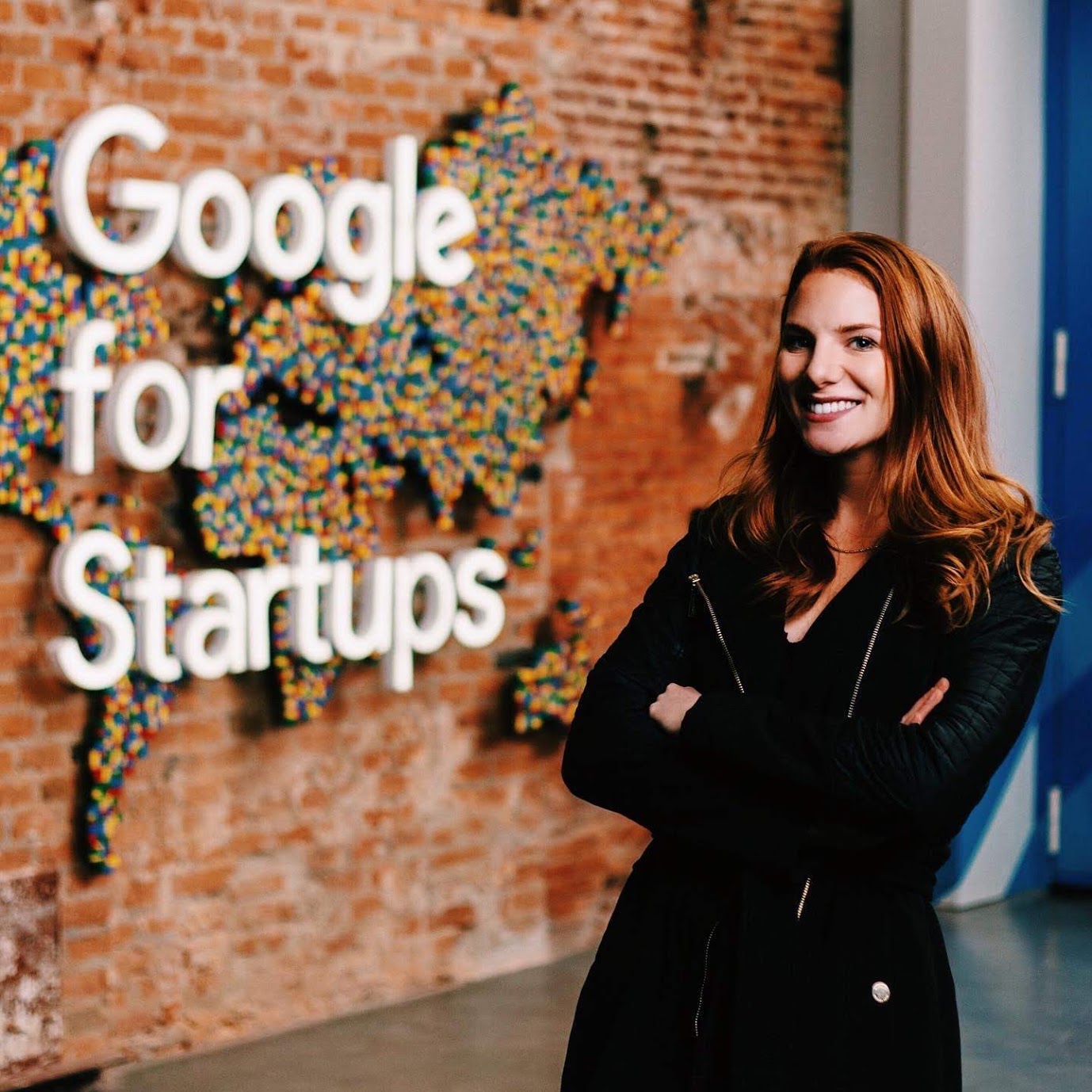 Photo of Hannah Frankl standing in front of a brick wall with a colorful map of the world and writing that says "Google for Startups". Hannah is a white woman with long read hair wearing a black shirt and jacket. She is smiling and posing with her arms crossed.