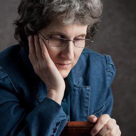 A headhsot of Kathi Wolfe. She is a white woman with grey hair. She is wearing a denim button down shirt and glasses. She is looking down with one hand on her cheek and the other on the back of the chair she shits on.