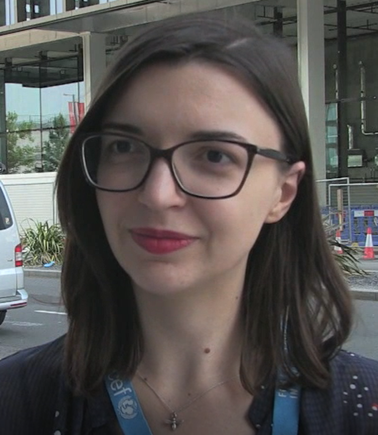 Pauline is a white woman with shoulder-length brown hair. She is wearing black glasses, red lipstick, and a black shirt. Behind her is a busy street.