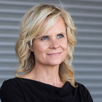 Headshot of Kristen Joiner. She is a white woman with curly blonde hair and blue eyes. She is wearing a black shirt and standing in front of a grey wall.