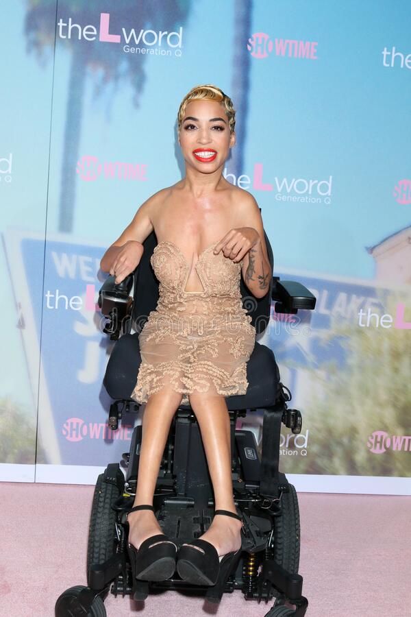 Jillian Mercado is a hispanic woman who uses a wheelchair and has short blonde hair. She is wearing a nude lace dress, black heels, and red lipstick. Behind her is a backdrop that read "Showtime" and "The L-Word: Generation Q"