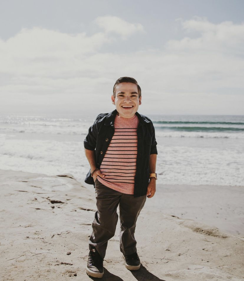 A photo of Brandon Farbstein standing on the beach in front of the ocean. He is a white, 22-year-old man who is 3' 9" and has brunette hair. He is wearing a pink shirt with black stripes, an open black button down, green pants, and black shoes.