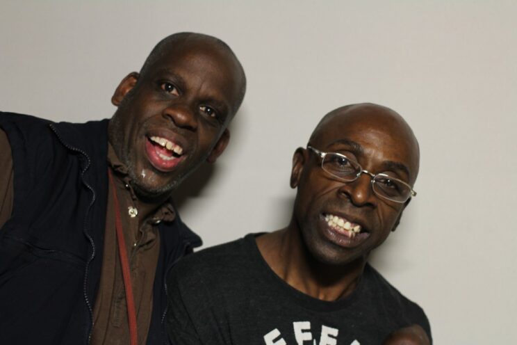 A close-up photo of Leroy Moore and Keith Jones standing together and smiling. Leroy, on the left, is a black man with gray close cropped hair and goatee. He is wearing a dark brown shirt with a black fleece vest. Keith, on the right, is a black man who is bald and is wearing wire glasses and a dark gray t-shirt.