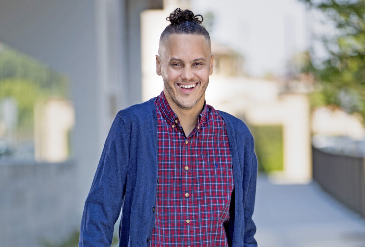 A headshot of James Ian, a black man with black hair shaved on the sides and the top tied into a man bun. He has light facial hair and is wearing a red and blue plaid button down shirt with a blue cardigan.