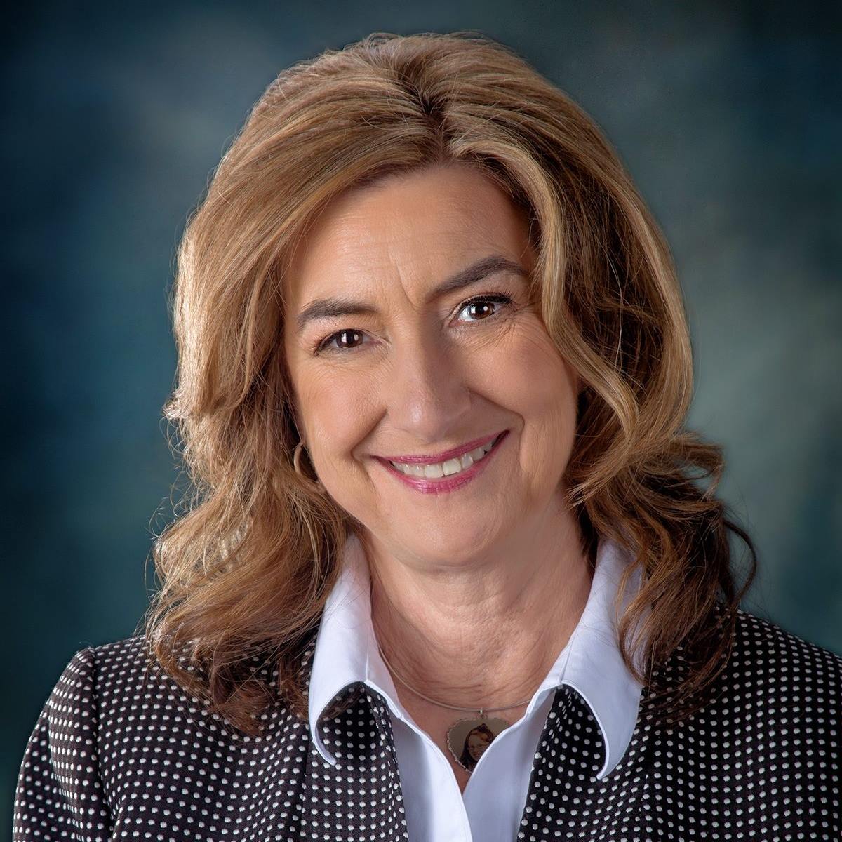 A headshot of Colleen Starkloff, a white woman with dark blonde hair. She is wearing pink lipstick, gold earrings, and a white collared shirt with a black and white polka dot blazer. She is smiling in front of a blue gradient background.