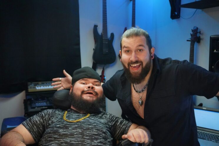 A photo of Zarek and Zack Elizondo in their studio. Zarek is a hispanic man who uses a wheelchair. He has a black beard and mustache. He is wearing a black hat, a grey shirt, and a gold chain. Zack is a hispanic man with brown hair pulled back into a bun and a brown beard. He is wearing a black button down that is open at the top with a two overlapping necklaces.