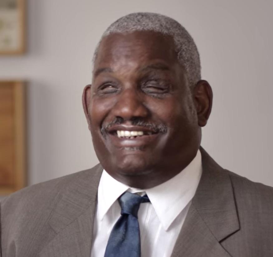 A photo of Dennis Billups smiling. Dennis is a Black blind man in his 60s with short grey hair. He is wearing a grey suit jacket with a white collared shirt and navy blue tie.