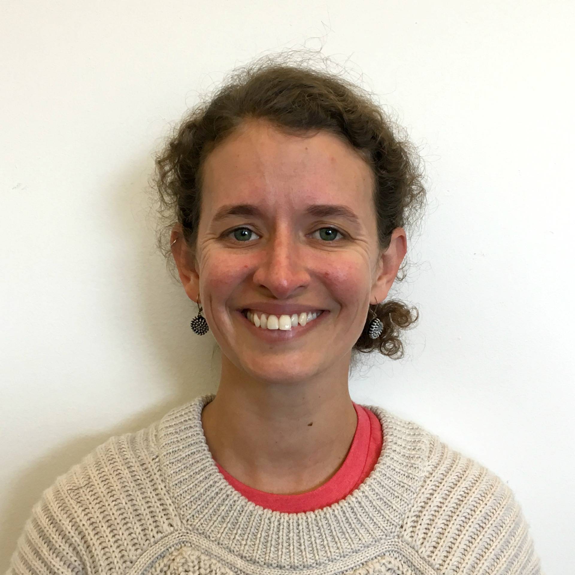 A photo of Emily Smith Beitiks smiling. Emily is a white woman with blue eyes and brown curly hair pulled back into a bun an eyes. She is wearing a cream sweater with a pink t-shirt underneath.