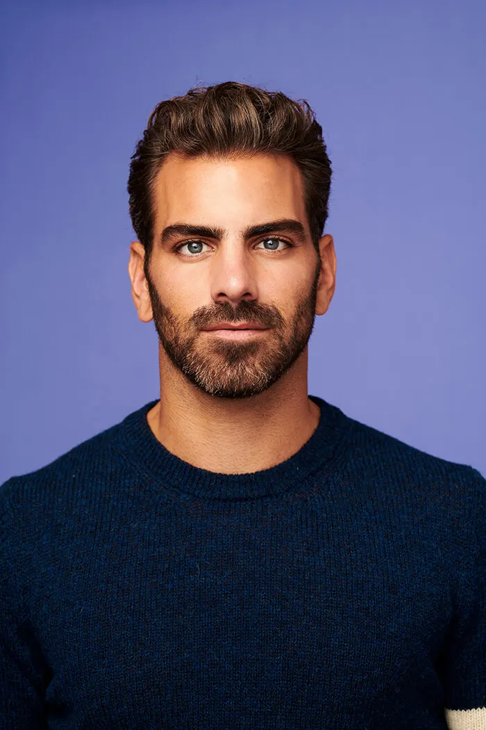 A headshot of Nyle DiMarco, a white man with dark brown short hair, light facial hair, and blue eyes. He is in front of a purple background wearing a dark blue sweater.