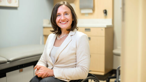 A photo of Cheri Blauwet, a white woman with short dark brown hair and blue eyes. She is wearing a white shirt, beige blazer, grey pants, and beaded necklace. She is sitting in her wheelchair in a doctor's office.