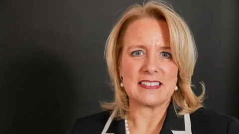 A headshot of Ann Cody, a white woman with shoulder-length blonde hair and blue eyes wearing red lipstick, a black blazer with white trimmed collar, pearl earrings and a pearl necklace.