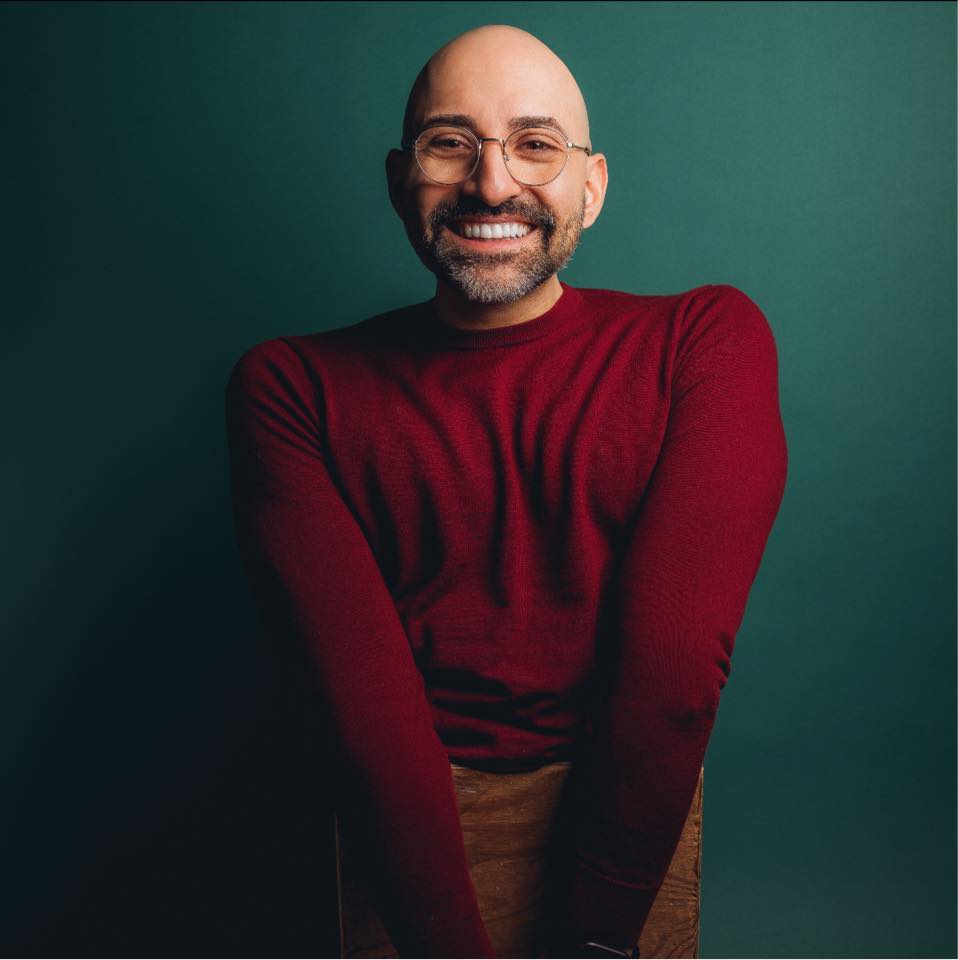 A photo of Spencer West, a white disabled man with no legs. He is wearing glasses and a red sweater. He is sitting on a brown box in front of a teal backdrop.