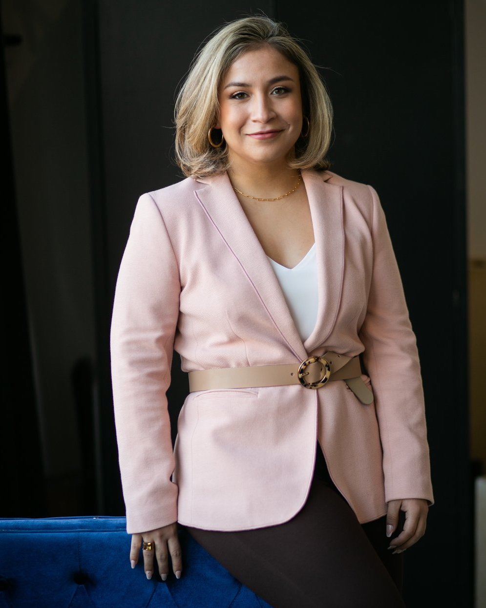 A photo of Dior Vargas, a light-skinned Latina woman with short blonde hair wearing black pants, a pink blazer with a white shirt underneath and a nude belt around her waist. She is leaning against a blue couch.