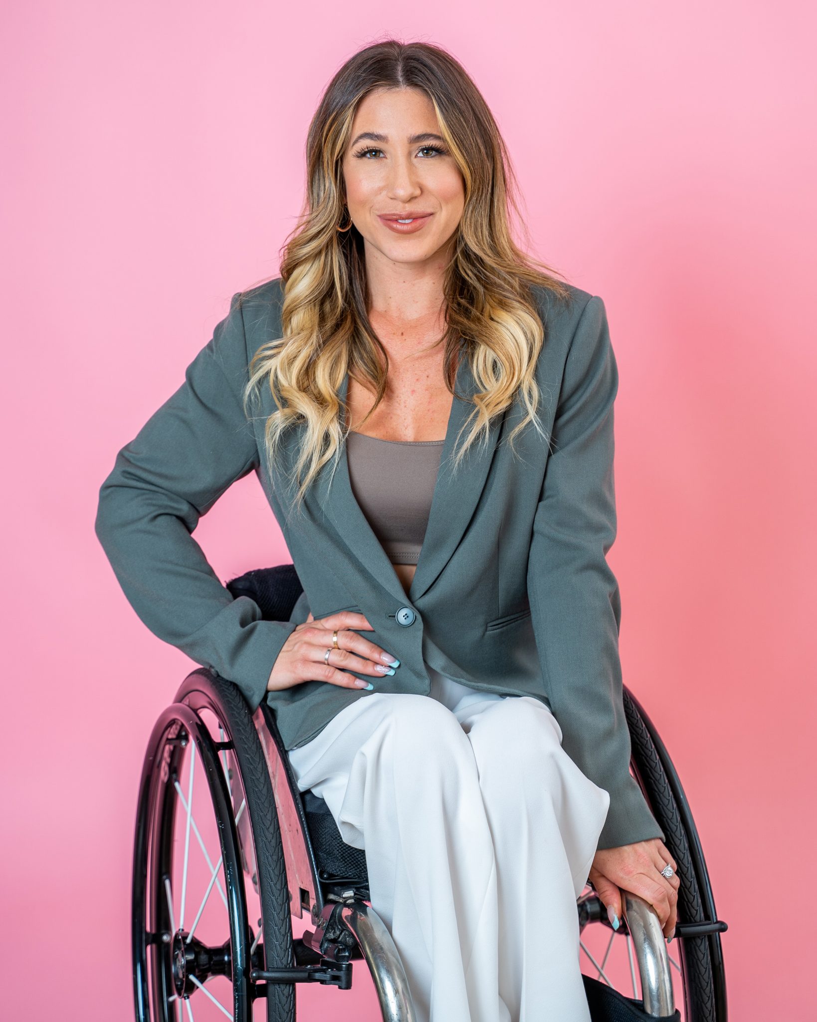 Photo of Chelsie Hill in front of a pink backdrop. She is a white woman using a manual wheelchair with long brown and blonde hair wearing a grey blazer and white pants.