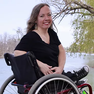 Photo of Dessa Cosma sitting in her wheelchair in front of a mural with different color pinks & blues in pattern. She smiles in a black tee shirt with blue dangling earrings. She has long blonde hair and light skin. Her arms come together in front of her lap with her fingers interlaced. 