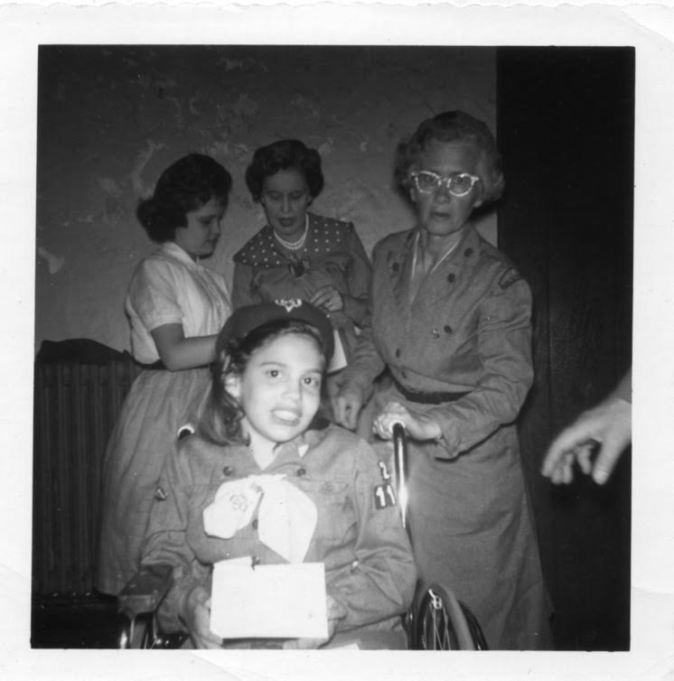 A black and white photo of Judy Heumann as a child in her Girl Scout uniform. She is white with brown hair and uses a wheelchair. Behind her are three women.
