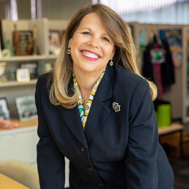 Headshot of Lidia Soto-Harmon, a Latina woman with shoulder length brown highlighted hair wearing a navy blazer with a girl scout pin.