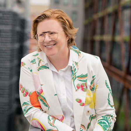 A photo of Lauren Ober, a white woman with short red hair wearing gold glasses, a white button down shirt, and a white blazer with colorful fruit designs on it.