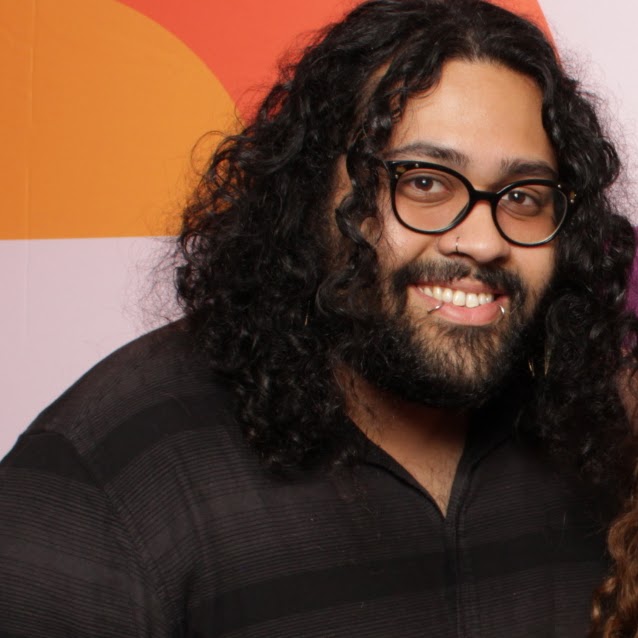 a photo of Vesper Moore a Brown, Hispanic, Mixed-Race, Indigenous person. They are wearing a black shirt with a stripe pattern, they are in front of an orange and peach circular pattern background.