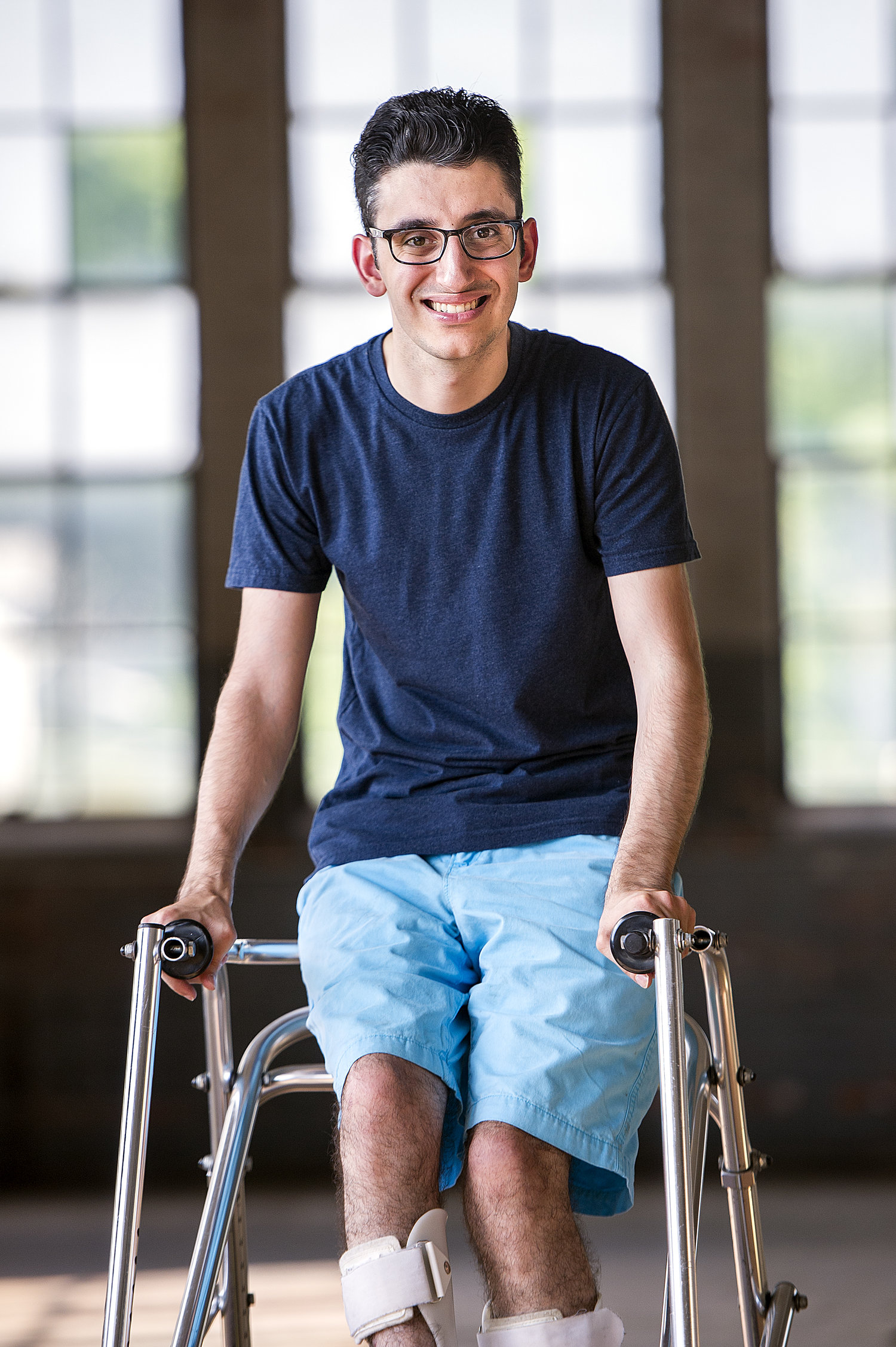 Ryan Haddad smiling with windows behind him. He is a Lebanese American man with short black hair and glasses who uses a walker. He is wearing a navy t-shirt and bright blue shorts.