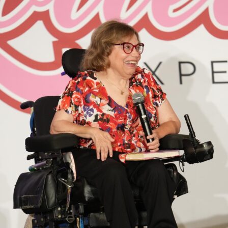 Judy Heumann holding a microphone on stage. She is a white woman wheelchair user with short brown hair wearing red glasses, a red floral blouse and black pants.