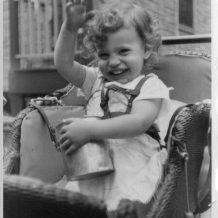 A black and white photo of Judy Heumann as a small child. She is wearing a dress and hitting a watering can while laughing.