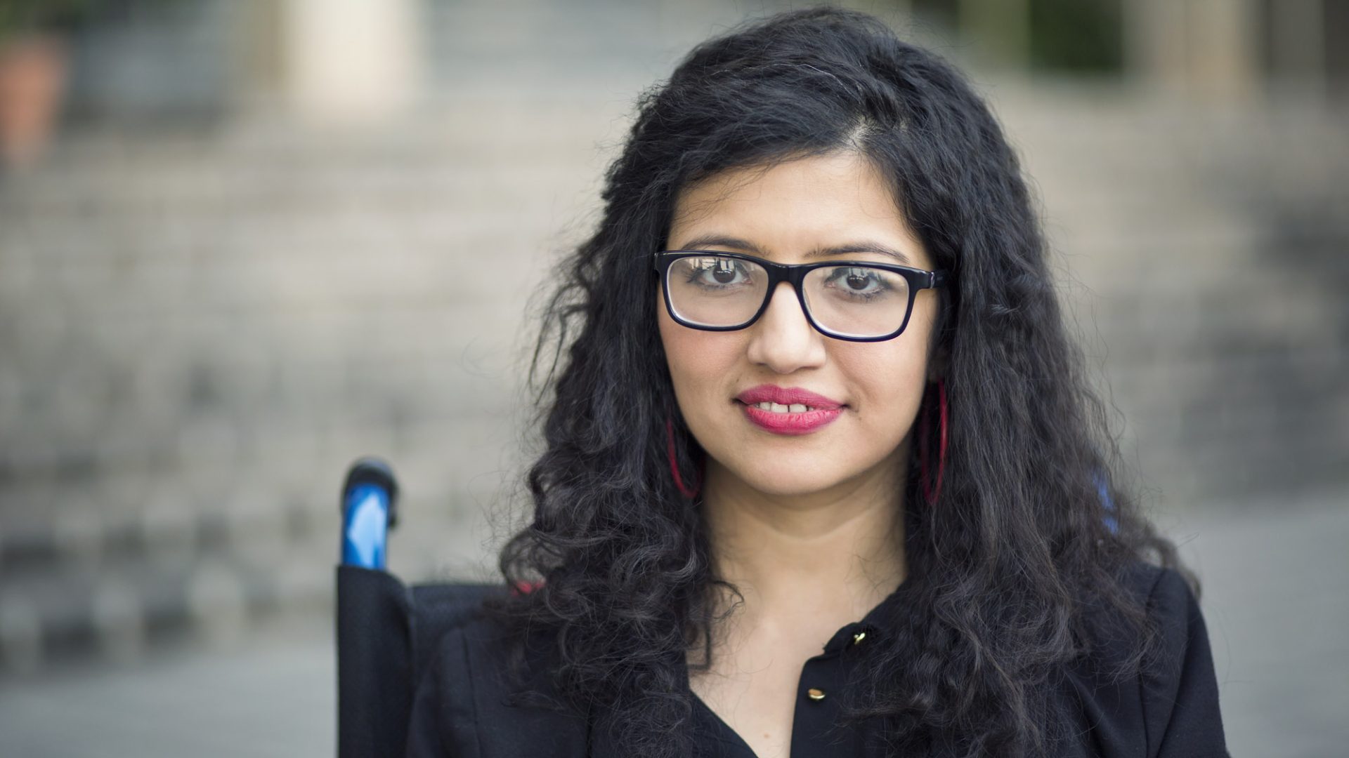 Headshot of Abia Akran, a Pakistani woman with long wavy black hair using a manual wheelchair and wearing glasses and a black shirt.