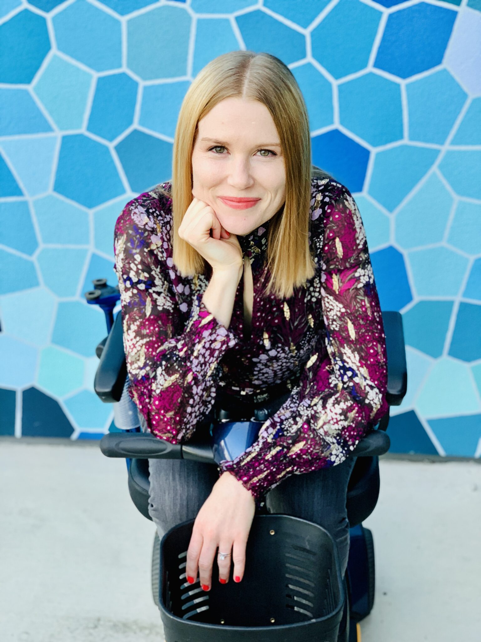 A blonde, white woman wearing a burgundy patterned shirt sits on a mobility scooter in front of a blue background with a variety of geometric shapes.