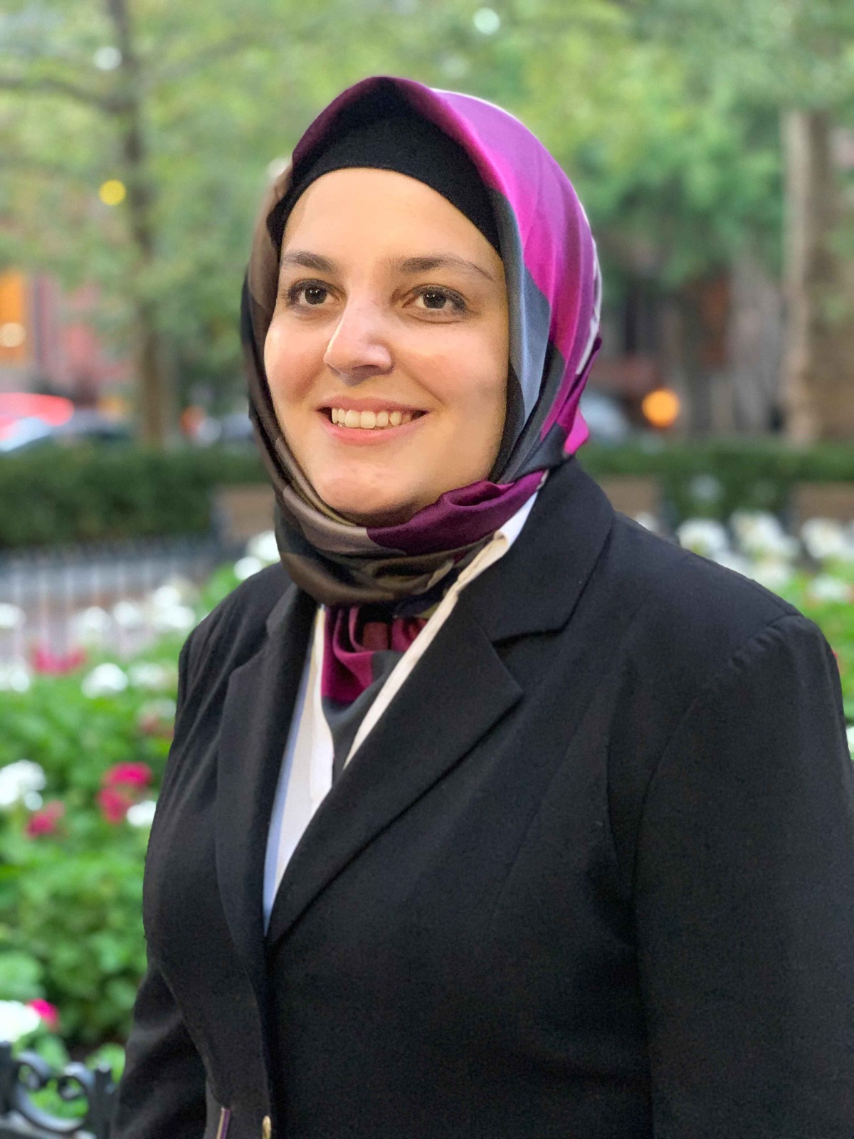 Mona Minkara, a Lebanese woman with light brown skin, wearing a purple hijab and black suit. Behind her are trees and flowers.