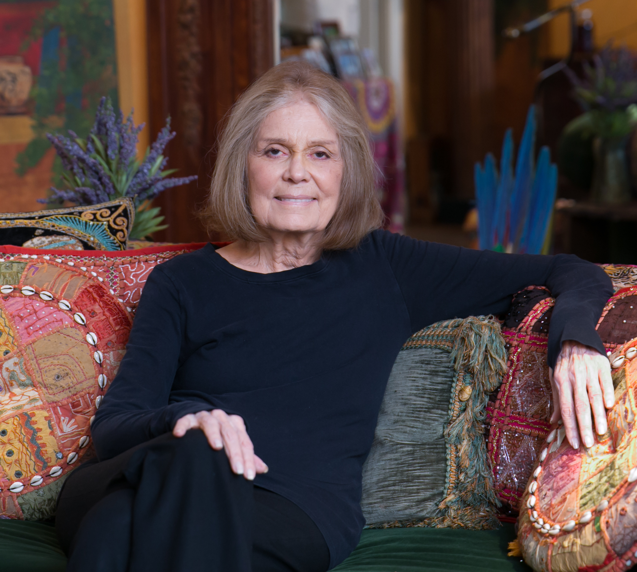 Gloria Steinem, a white woman with dark blonde and grey hair wearing a black shirt and black pants seated on a colorful chair.